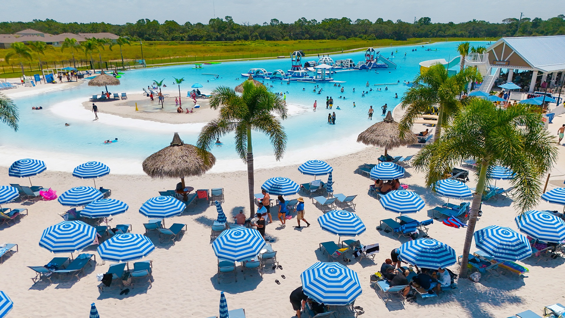 Aerial view of a Crystal Lagoons Public Access Lagoon with inflatable water park and beachgoers under striped umbrellas
