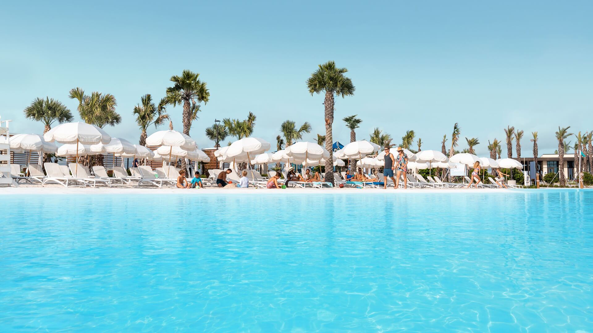White beach umbrellas and palm trees along the sandy shore of a crystalline lagoon