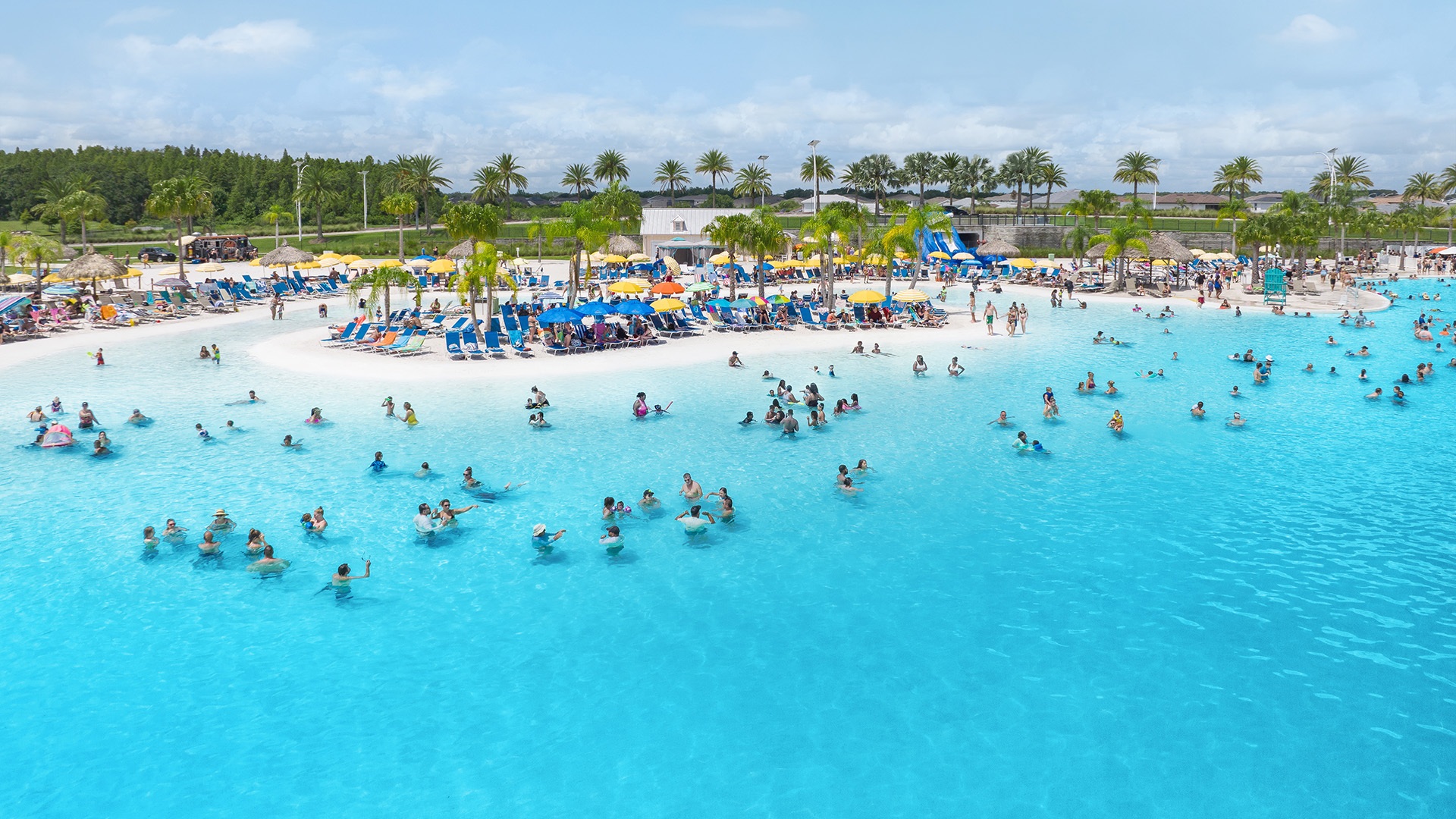Aerial view of a crowded Public Access Lagoon with swimmers, white sand beach and colorful umbrellas